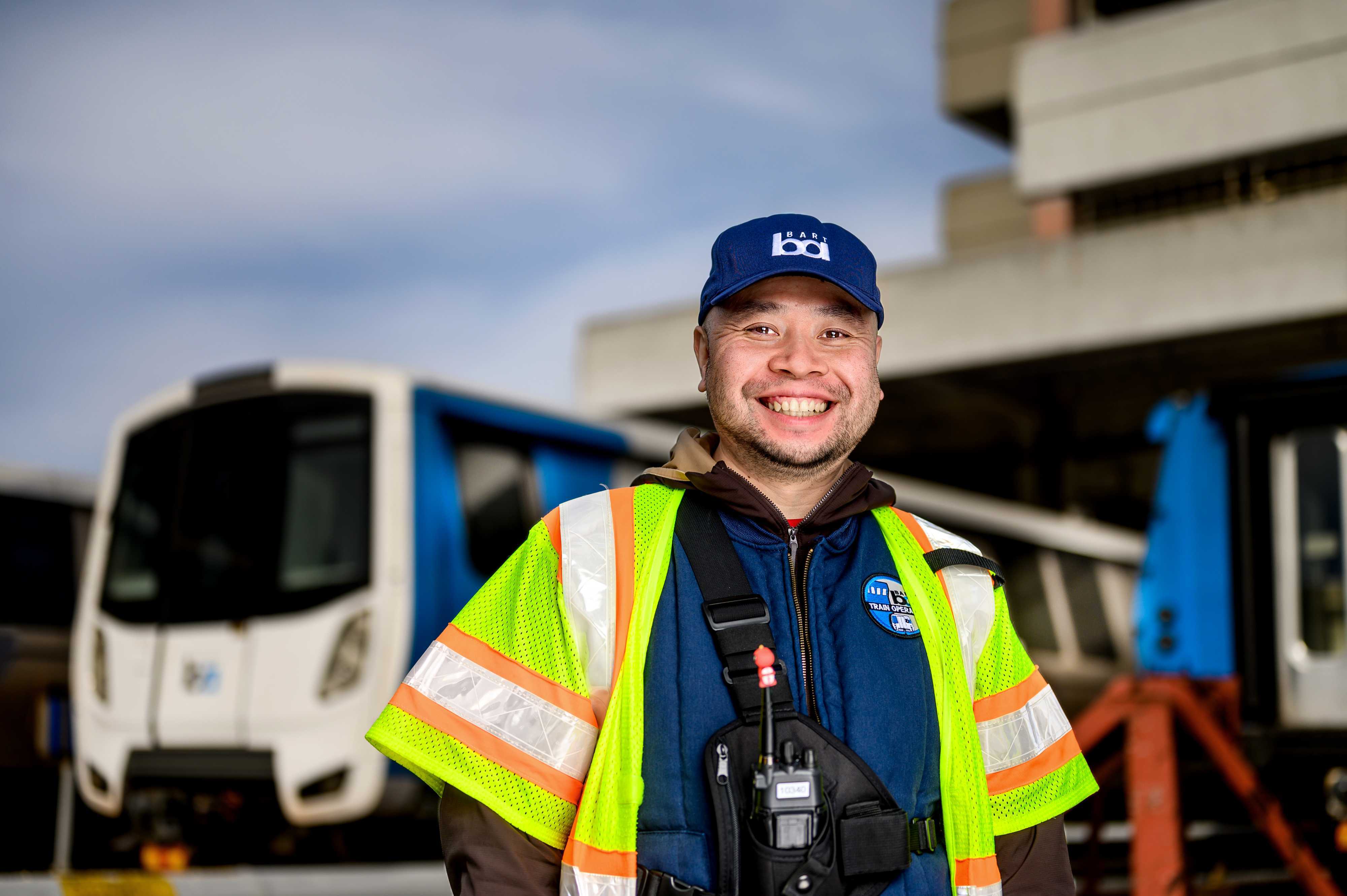 A person in a reflective vest and BART hat stands in front of a bart train.