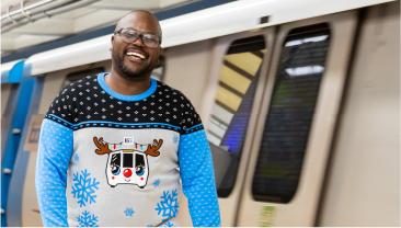 Person smiling in a BART holiday sweater in front of a BART train 