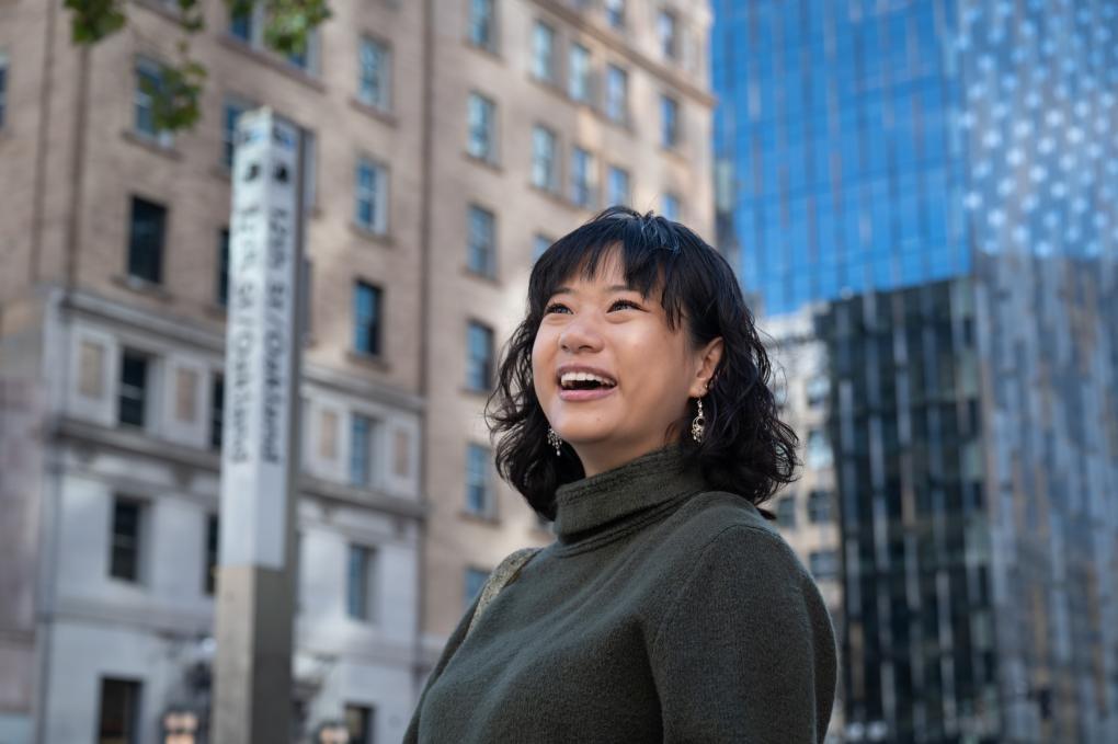 A woman in a green turtleneck smiles in front of buildings by 12th St