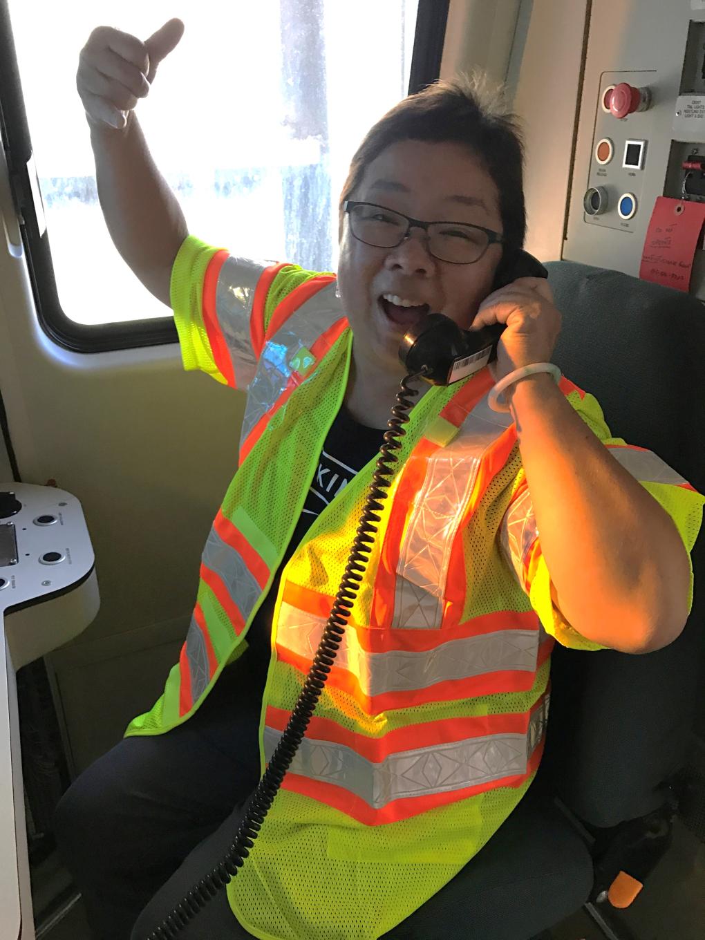 A recent photo of Mama Linda in a train car cab in a yellow vest doing a thumbs up and holding a black phone,.