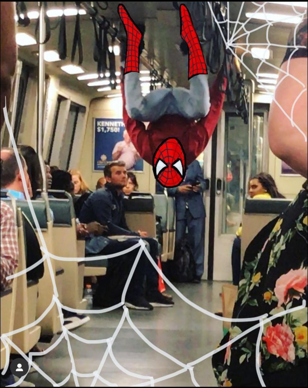 An individual dressed in a Spider-Man costume is performing a handstand on a moving train, with their feet raised towards the ceiling. The train interior is populated with various passengers seated and standing.
