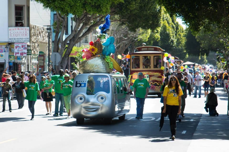 A lively street parade featuring a colorful float designed like a cartoonish blue train with large eyes and a decorative peacock on top. The float is surrounded by people in green outfits, and other decorated vehicles are visible in the background. There are bystanders and buildings lining the sunny street.