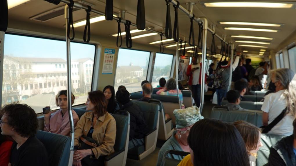 Interior of a legacy train with passengers seated and standing, holding onto overhead handles. Windows show buildings outside.