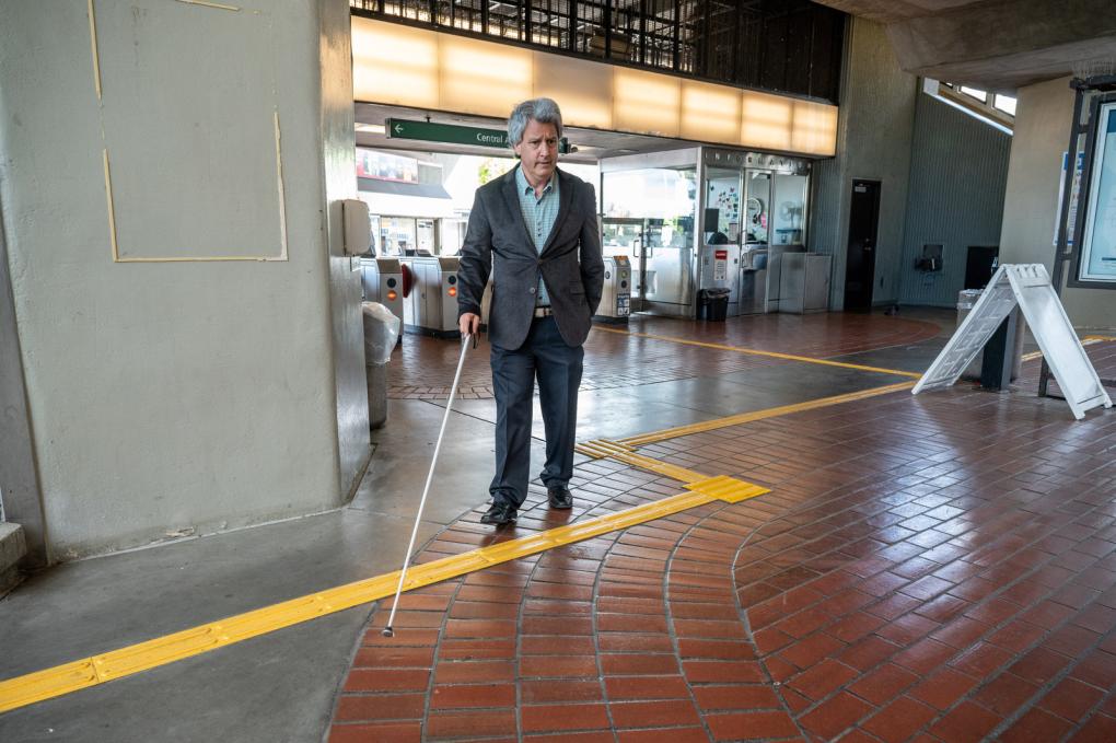 A person using a white cane is navigating through a BART  station. They are walking on a tactile paving designed for visually impaired individuals. The background shows ticket machines and a gated entryway. The setting includes ample natural light with architectural details like beams and columns.