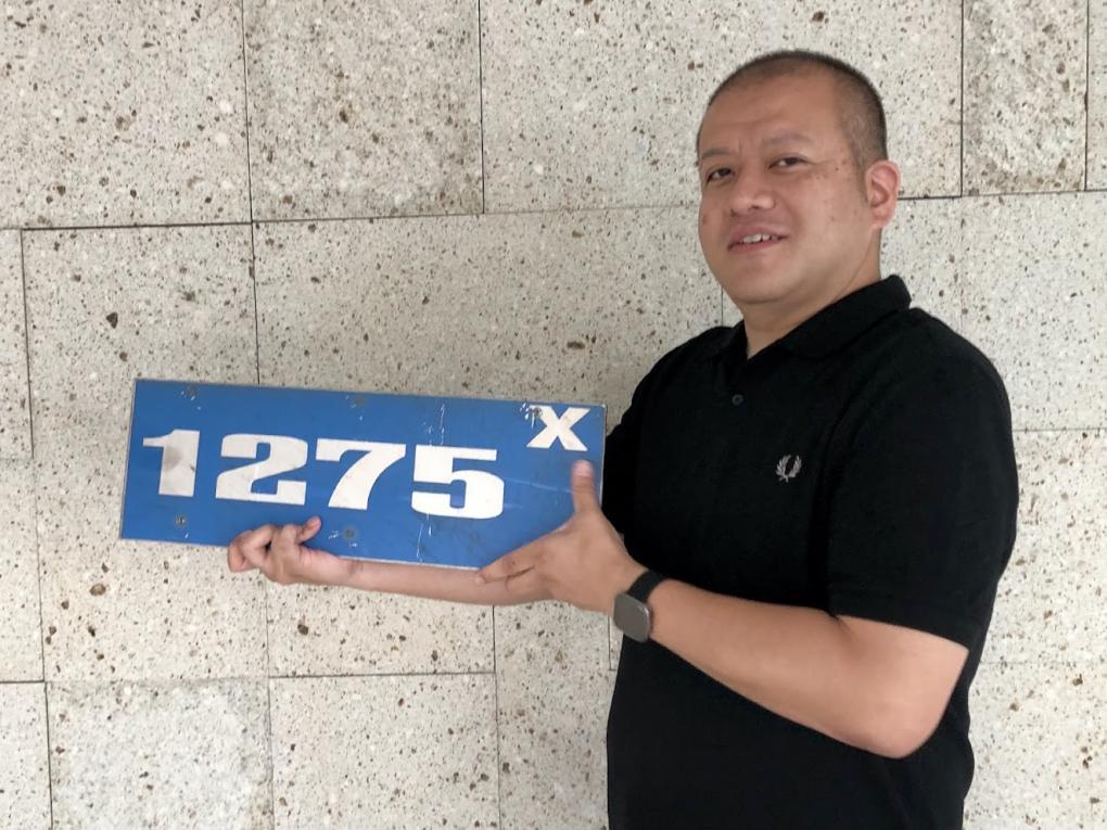 Atsushi holding a blue number plate with the number 1275X, smiling, standing against a speckled beige wall.