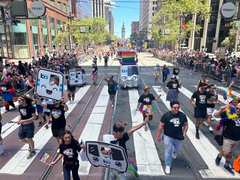 A vibrant Pride parade in San Francisco, featuring participants carrying square cutouts of train faces. A decorated BARTmobile with a large rainbow hat is also visible. The scene is bustling with onlookers lining the streets.