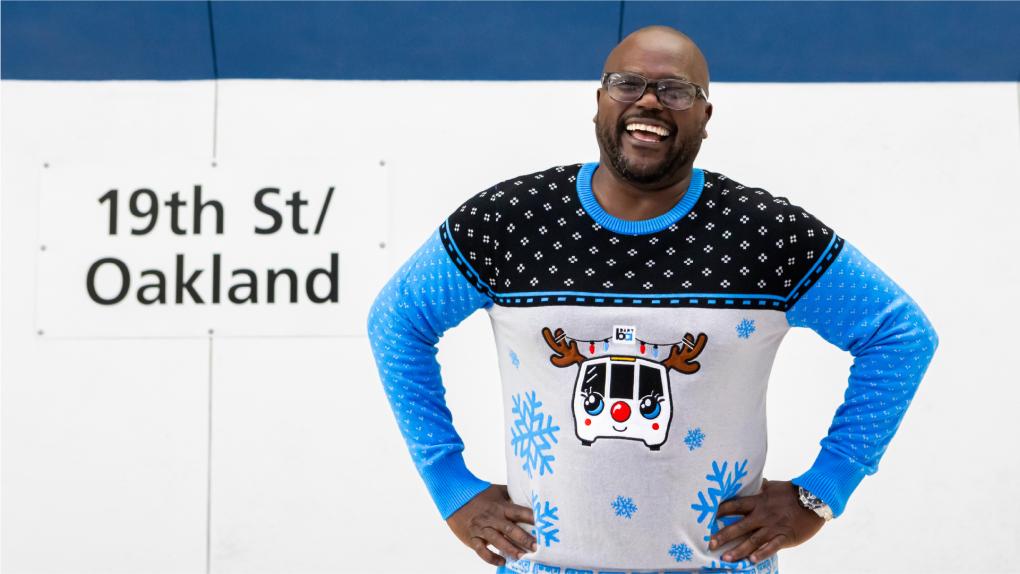 Person smiling in a BART holiday sweater in front of the words 19th St/Oakland