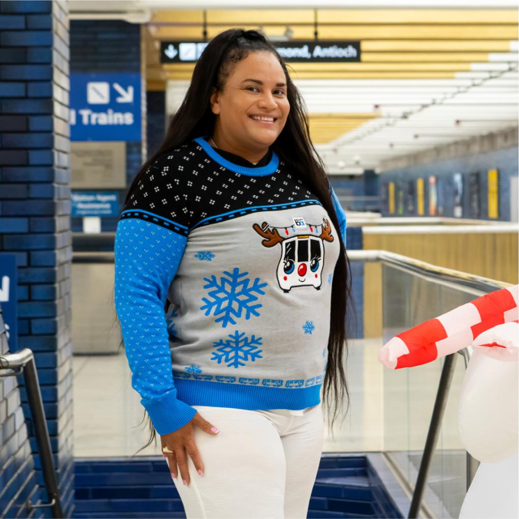 Person smiling in a BART holiday sweater in front of a BART train 