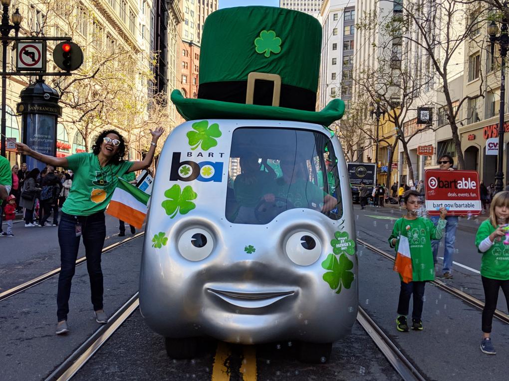 The BARTmobile decorated with a face and green shamrock motifs, including a large, green Irish-themed hat, participates in a parade on a city street. People, including a person waving enthusiastically, walk beside it celebrating.