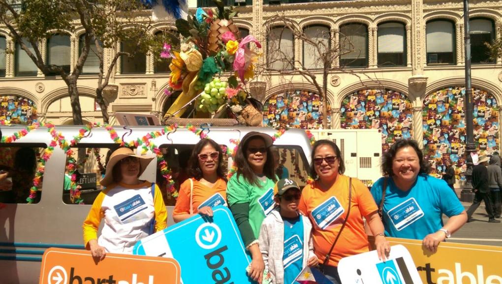 A group of people wearing hats and sunglasses participating in a street parade. They are standing next to a festively decorated float featuring a large floral arrangement. Behind them, a historic building with intricate architectural details is visible.