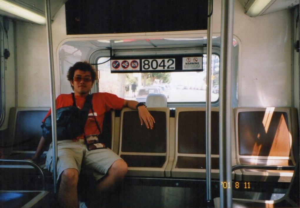 A person sitting in a bus, comfortably sprawled across two seats. They are wearing sunglasses, a red shirt, and are holding onto a bag strapped across their body. The bus interior is visible with empty seats and signs above the windows showing various symbols, including no smoking and a wheelchair access symbol. The bus number "8042" is displayed at the front. The image also includes the date "01 8 11" in the lower right corner.