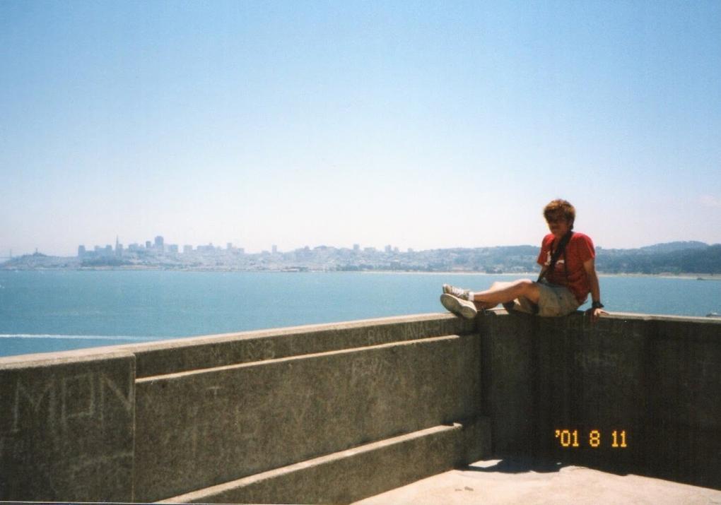 Atsushi sits on a concrete ledge with a panoramic view of the San Francisco skyline and bay in the background, on a sunny day.