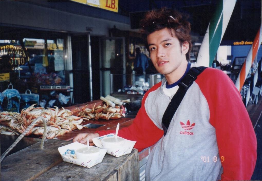Atsushi sitting at a seafood stall with multiple crabs on display in front of them, wearing a red Adidas sweatshirt and a black shoulder bag, enjoying a meal from two small bowls. Visible date stamp "01 8 9.