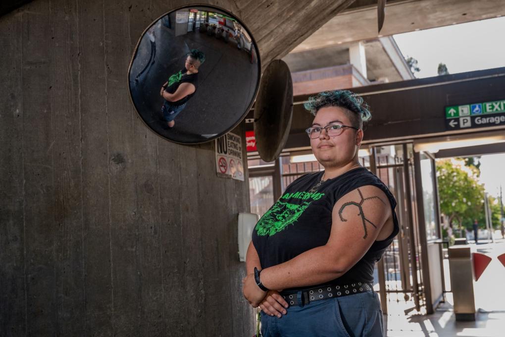 Sam Johnson smiling in a station with their visible tattoo. There is a reflectin in the security mirror.