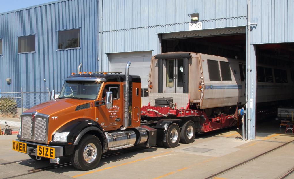 A large, orange truck marked "OVER SIZE" pulling a trailer with a BART car, parked beside a blue industrial building with open doors.