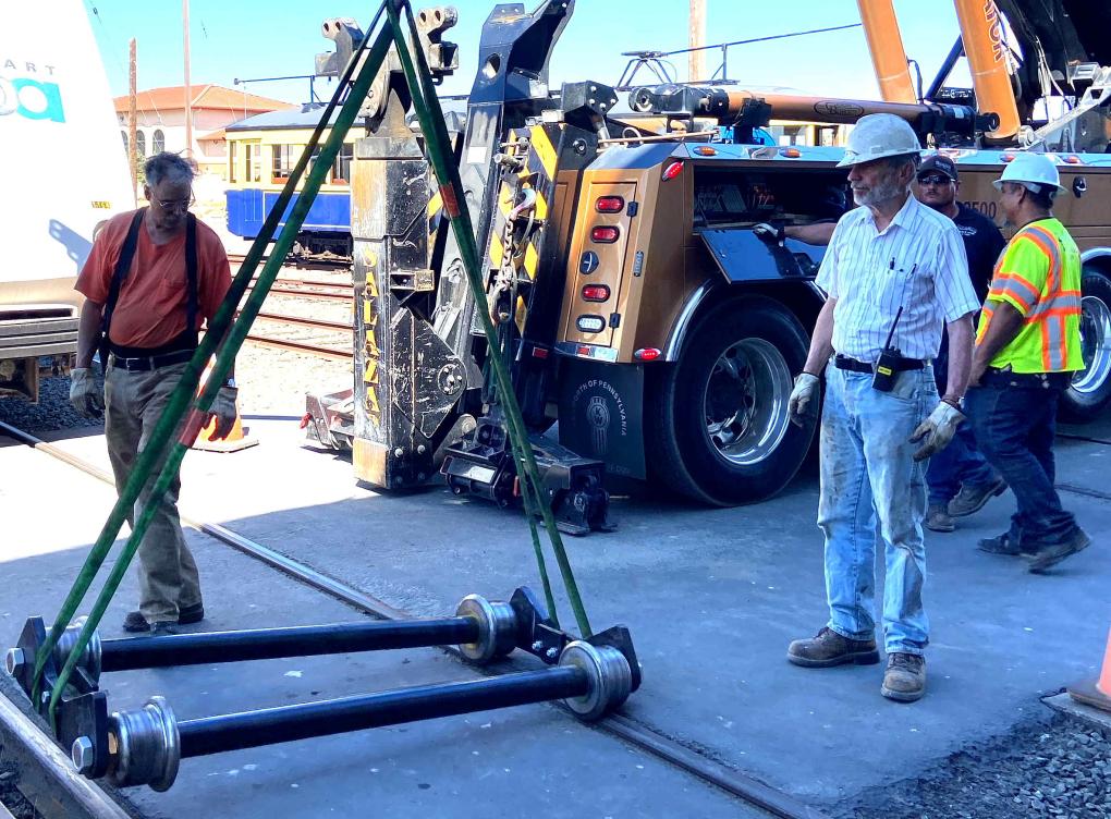 Three workers with heavy machinery, including a crane and a tow truck, in the background. One worker is walking past a large metal frame on the ground, while the other two are standing and conversing.