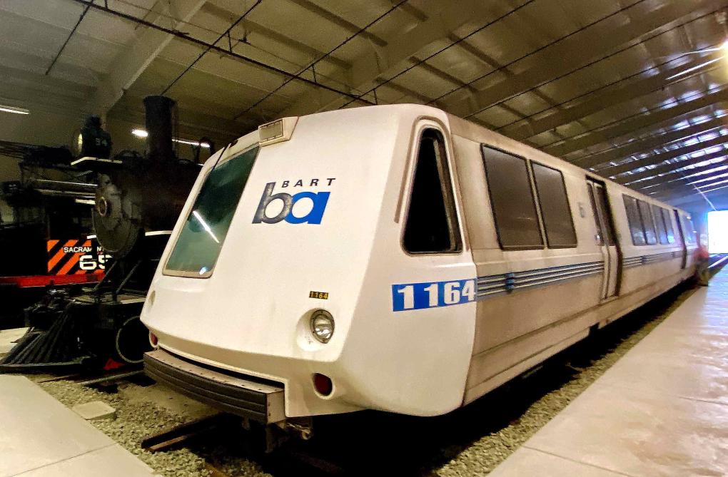 A BART car parked in a warehouse, with an older locomotive visible in the background.