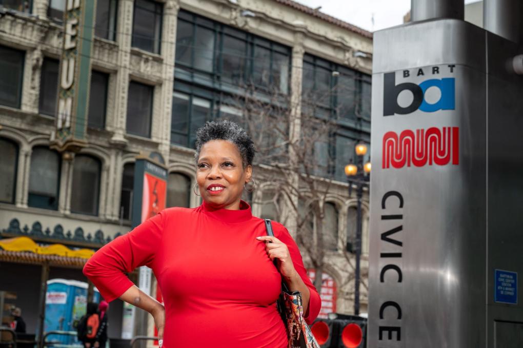 Sherri Young pictured next to a Civic Center BART and Muni sign in front of the Orpheum Theater. She is wearing a red dress and carries a purse