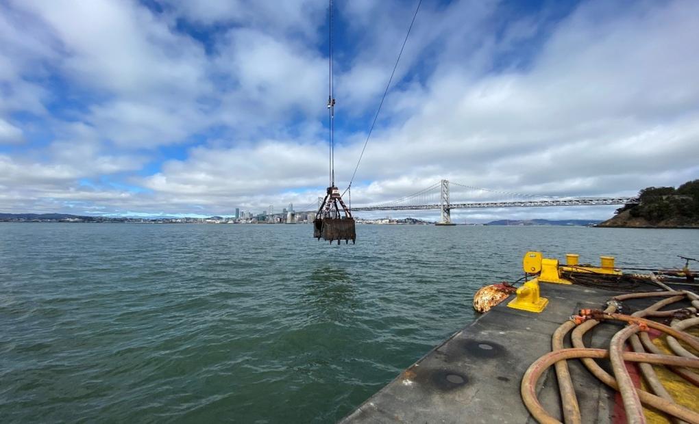 Large clamshell dredging bucket being lowered into the bay as part of soil handling operations.