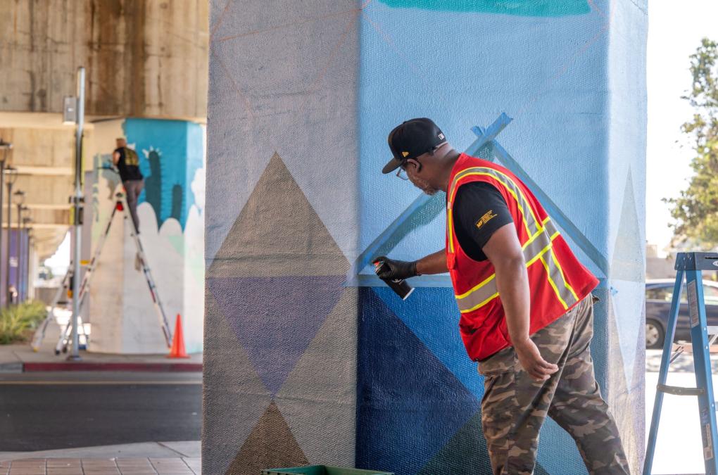 A person in a reflective vest and cap spray painting a geometric mural on an urban wall, while another person in the background works on a mural.