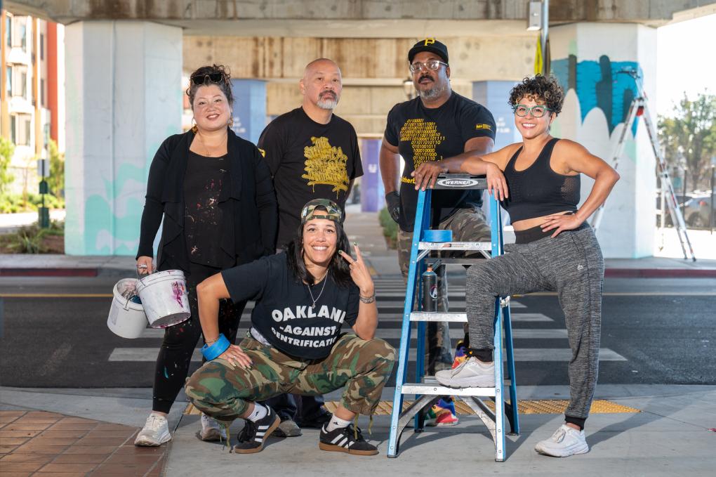 Five artists pose with paint supplies under the platform at Fruitvale Station, featuring a mural in progress. Two individuals are standing, two are kneeling, and one is seated on a ladder. 