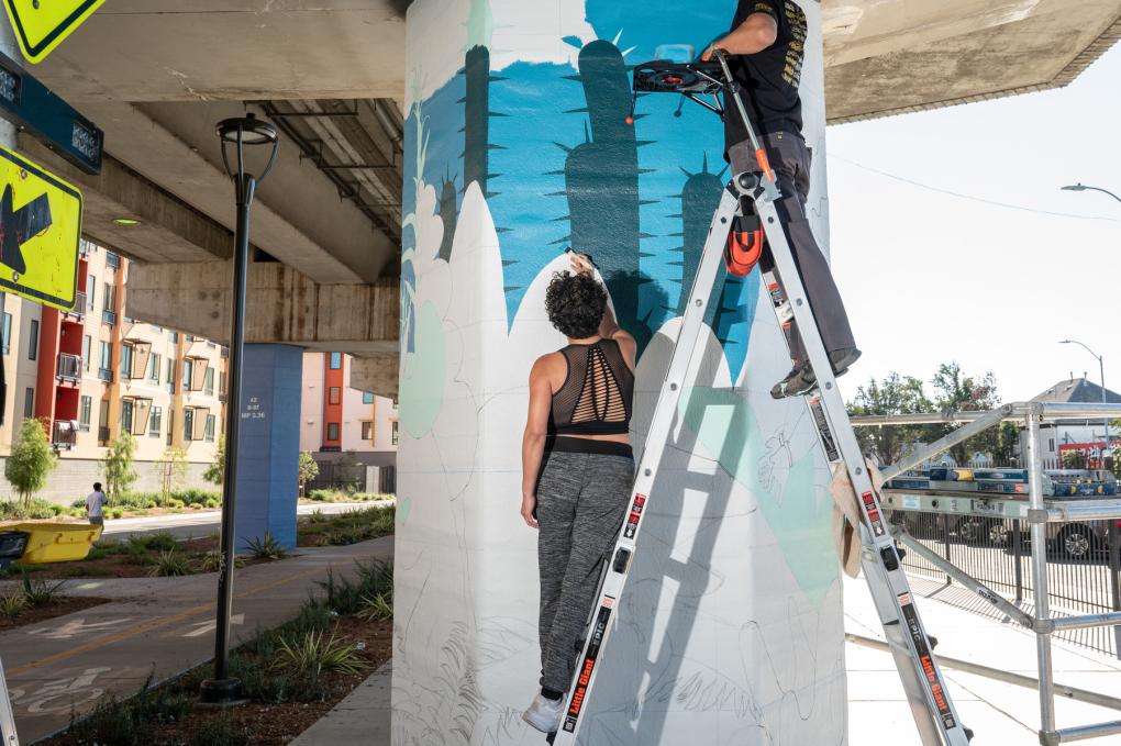 Favianna Rodriguez and Vogue paint an unfinished mural on a column under Fruitvale Station platform.