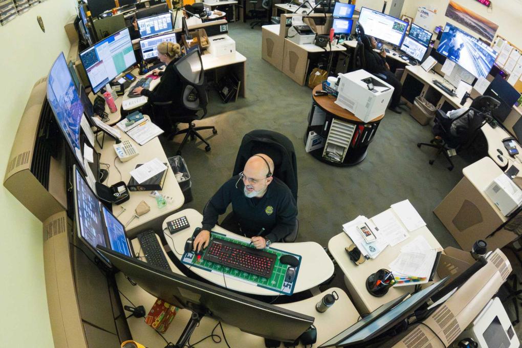 A dispatcher smiles at his desk in the dispatch center with lots of computer screens and others working