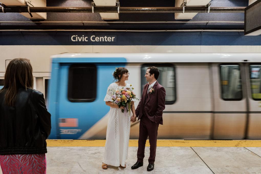 Couple in wedding attire smile on a bart platform